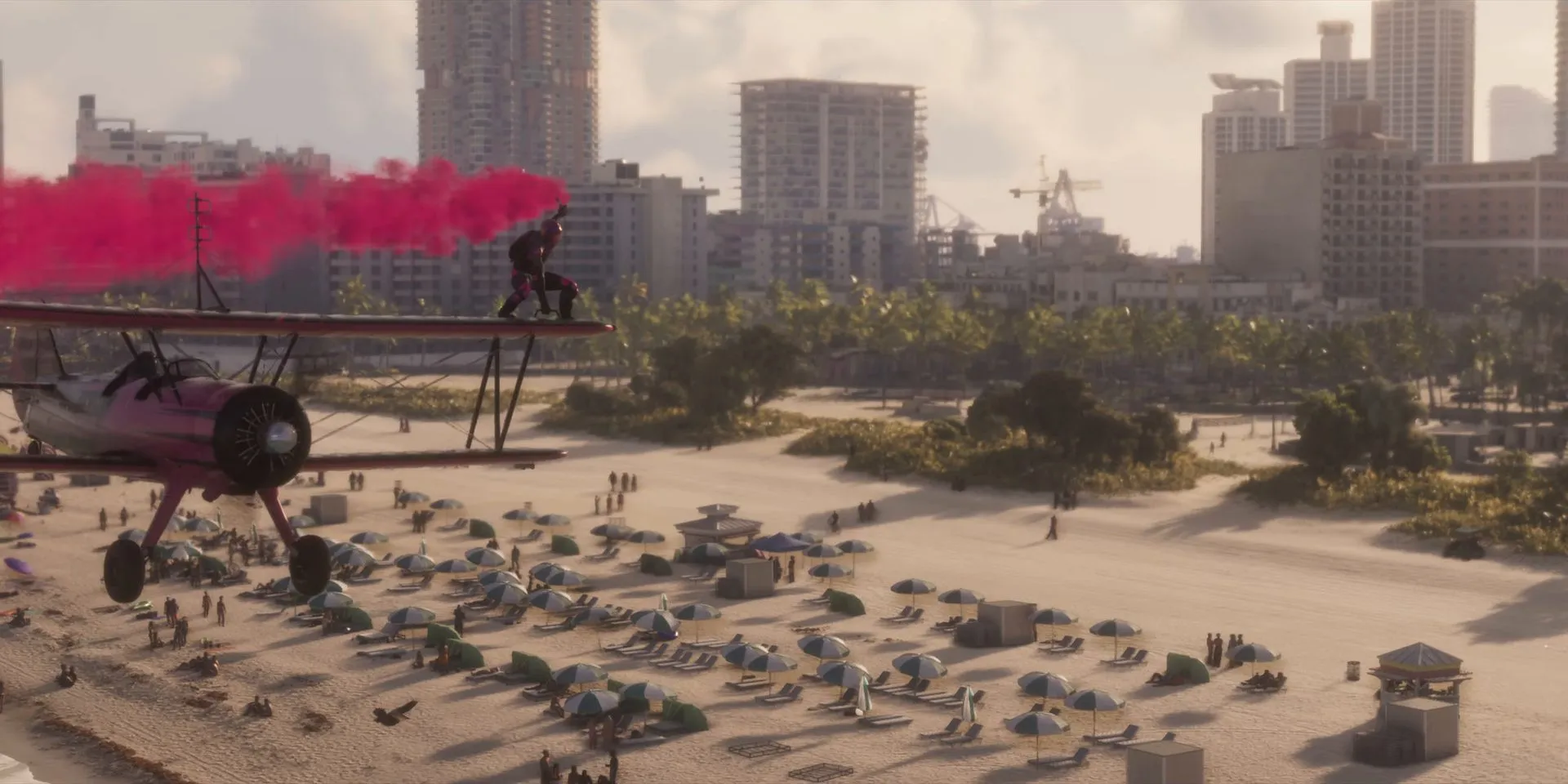 A pink biplane trails pink smoke low over a crowded beach with high-rises behind